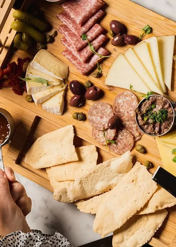 A person's hand serves a wooden charcuterie board with cheese, meats, crackers.