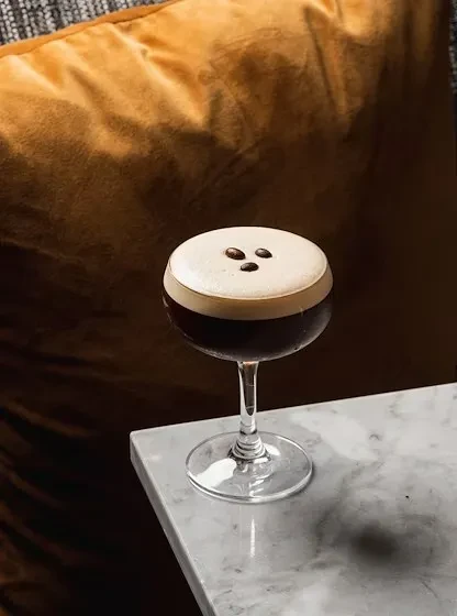 Dark cocktail with creamy foam and coffee beans in a coupe glass.