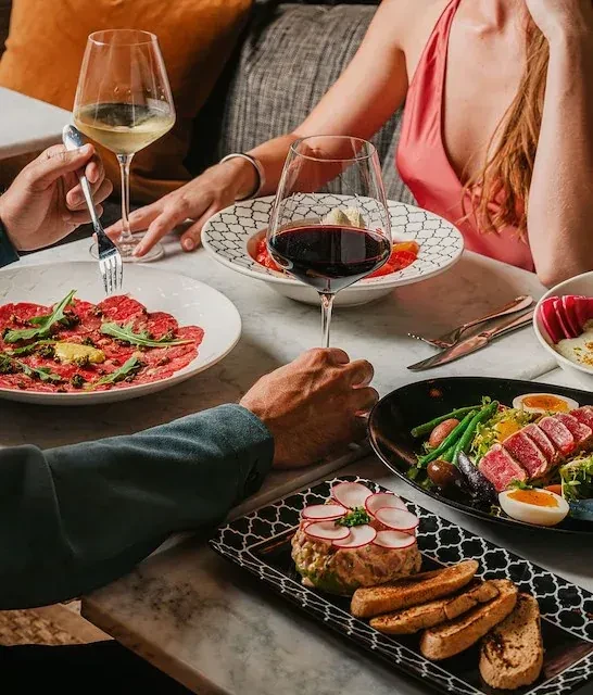 Restaurant table with carpaccio, tuna, tartare, wine, and diners' hands.