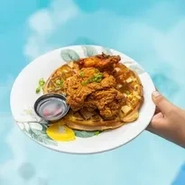 A hand holds a plate of golden chicken and waffles with syrup.