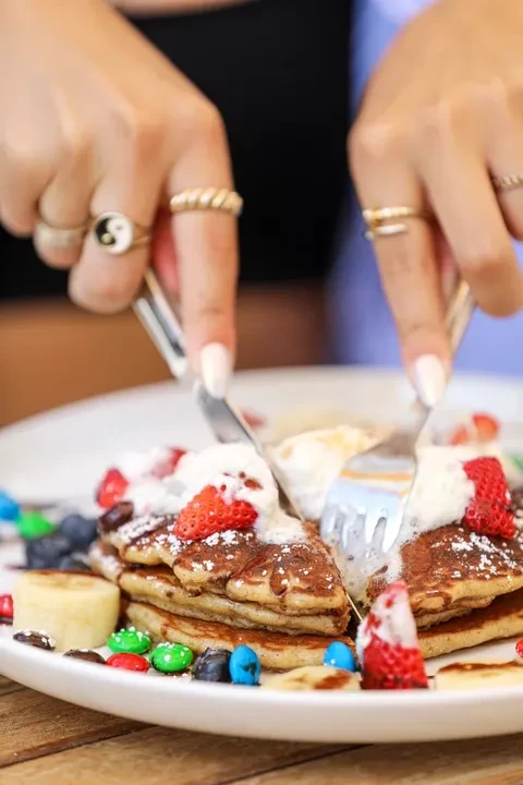 Hands cutting pancakes with strawberries, whipped cream, assorted fruits, and candy.
