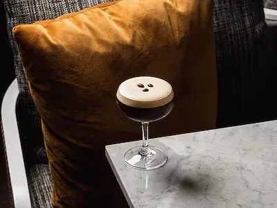 Dark cocktail with white foam and coffee beans on a marble table.
