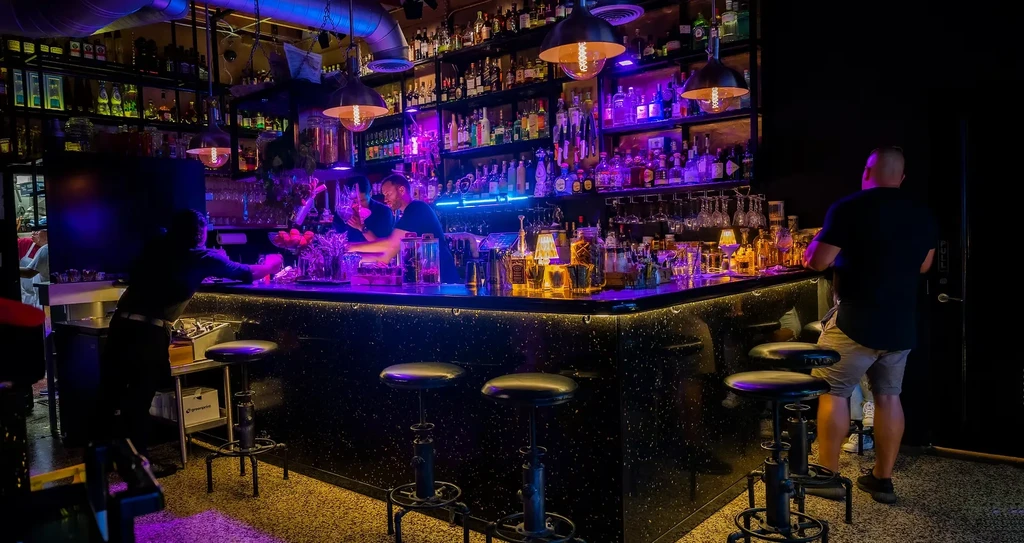 Bartender preparing drinks at a well-stocked bar with vibrant neon lights.