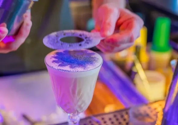Bartender sprinkles purple powder through stencil onto frothy cocktail.