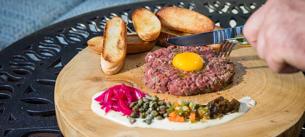 Beef tartare with egg yolk, garnishes, and baguette on wooden board.