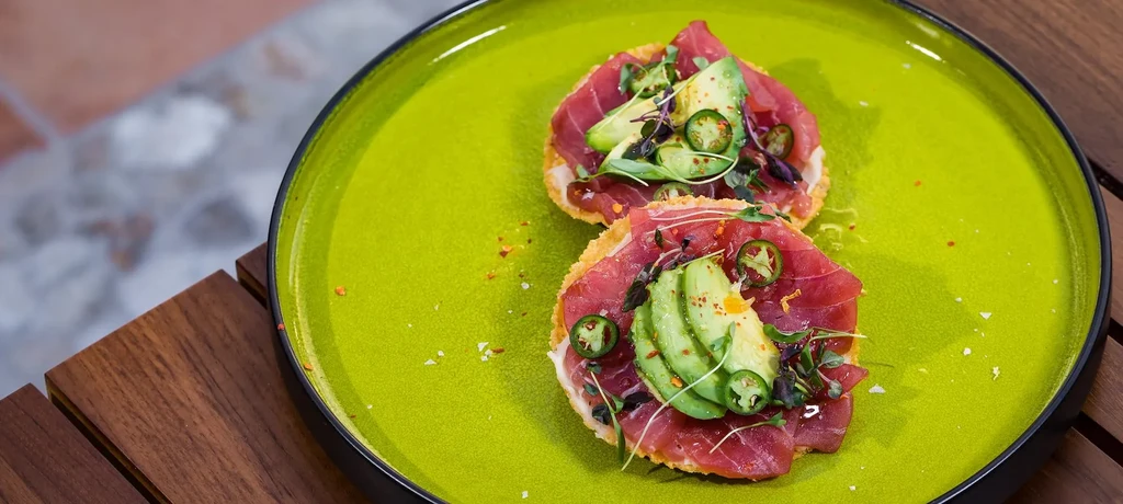 Two tostadas with raw fish, avocado, jalapeño, microgreens on a green plate.