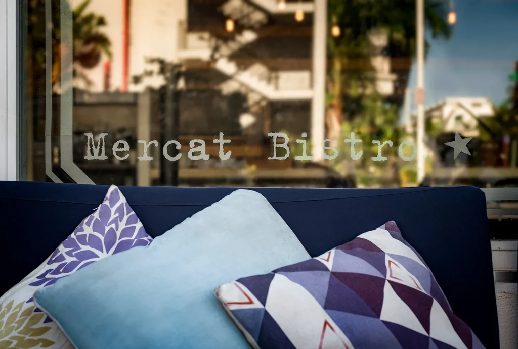 Three decorative pillows on a navy sofa, 'Mercat Bistro' on blurred window.