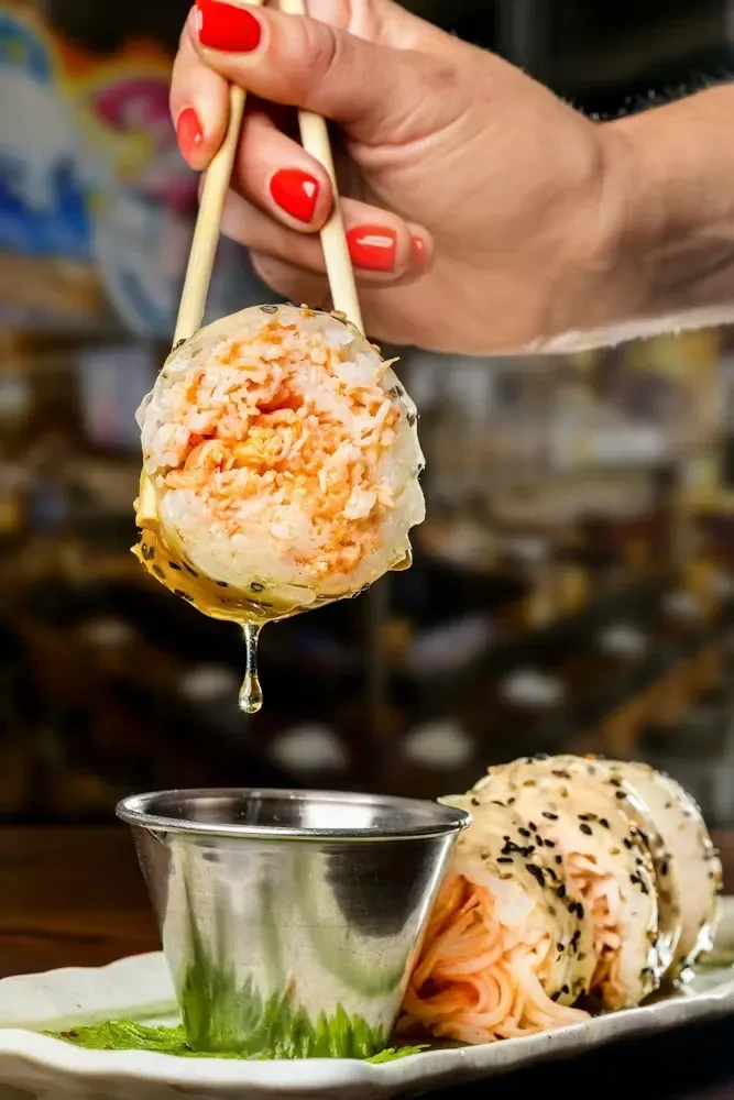Chopsticks hold orange-filled sushi roll, sauce dripping into a cup.