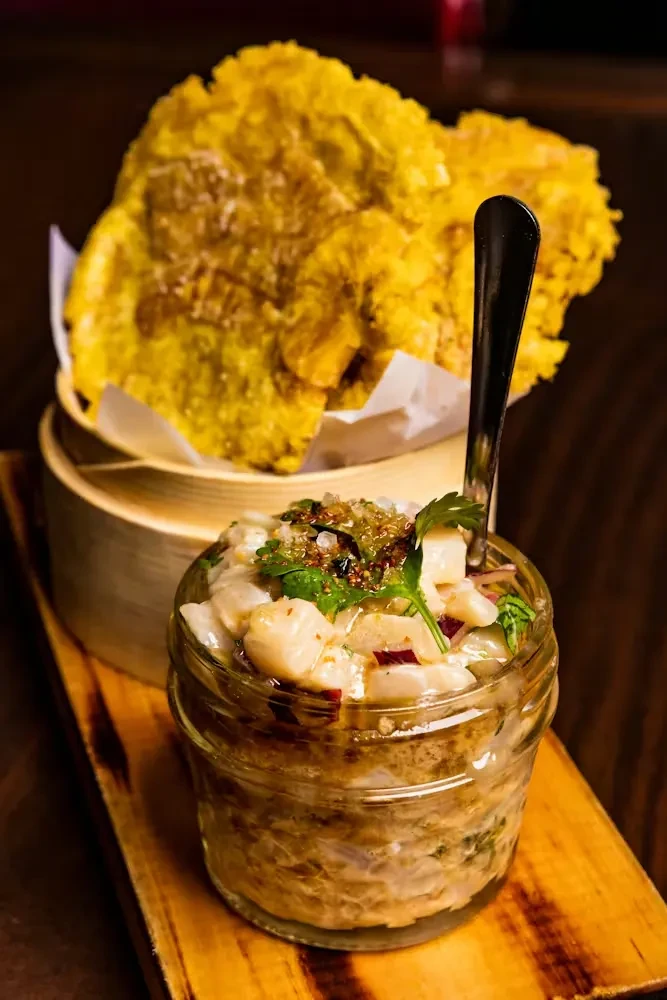 Ceviche in glass jar, crispy plantains in steamer, on wood.