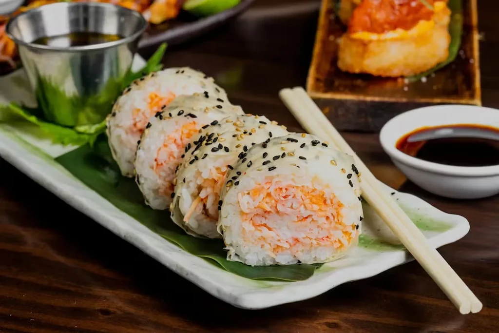 Plate of four sesame sushi rolls with soy sauce and chopsticks.