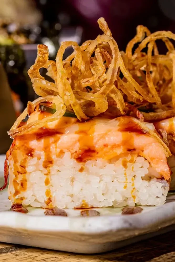 Close-up sushi roll with shrimp, drizzled sauces, and crispy onions.