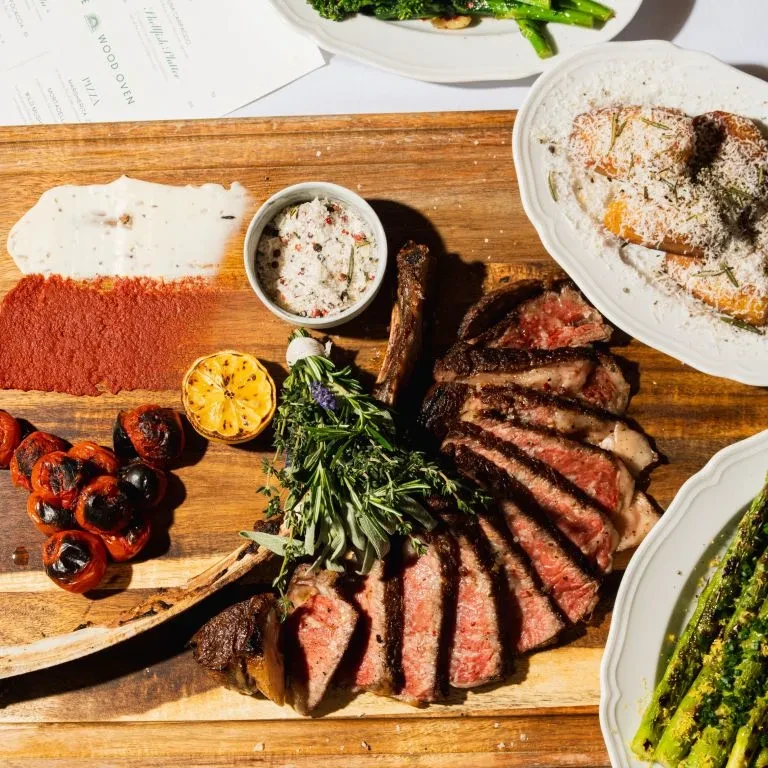 Sliced tomahawk steak, medium-rare, with sauces, herbs, and plated sides.