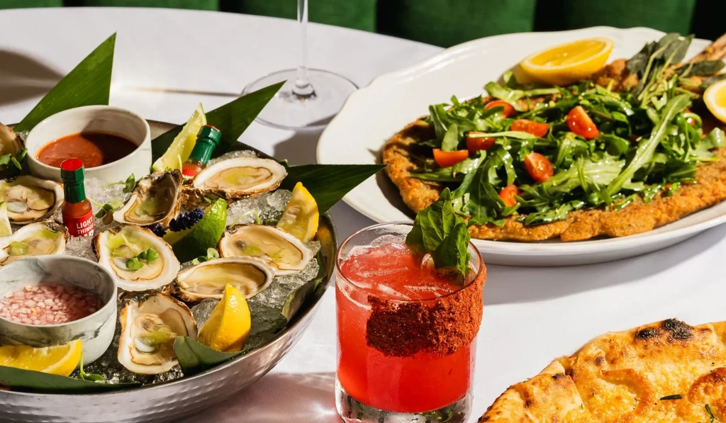 Well-composed table with oysters, breaded cutlet with salad, red cocktail, and flatbread.