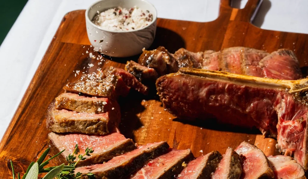 Sliced medium-rare steak on a wooden board, being salted, with herbs.
