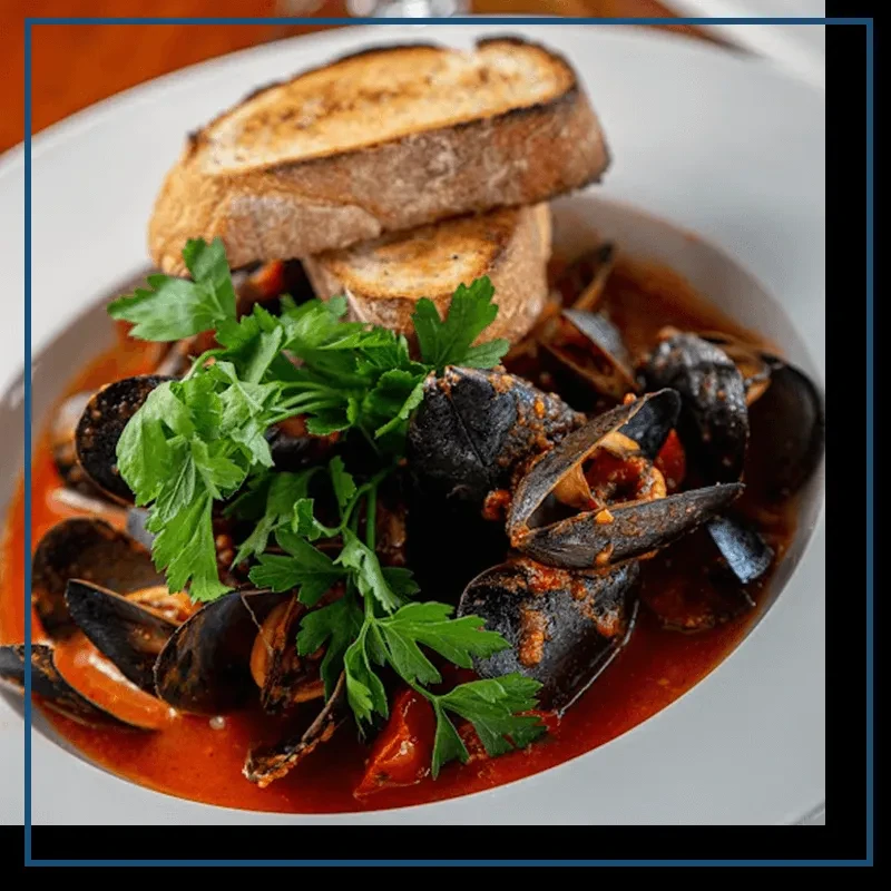 Mussels in tomato broth with bread and parsley in a white bowl.