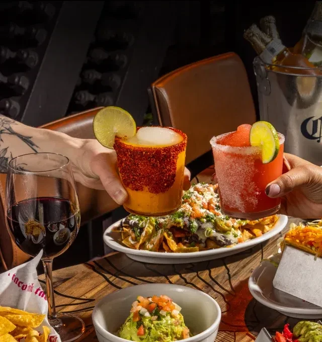 Hands toast with cocktails over nachos, guacamole, and a Mexican spread.