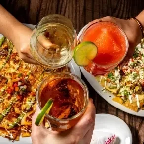 Three hands toast drinks over nachos on a dark wooden table.