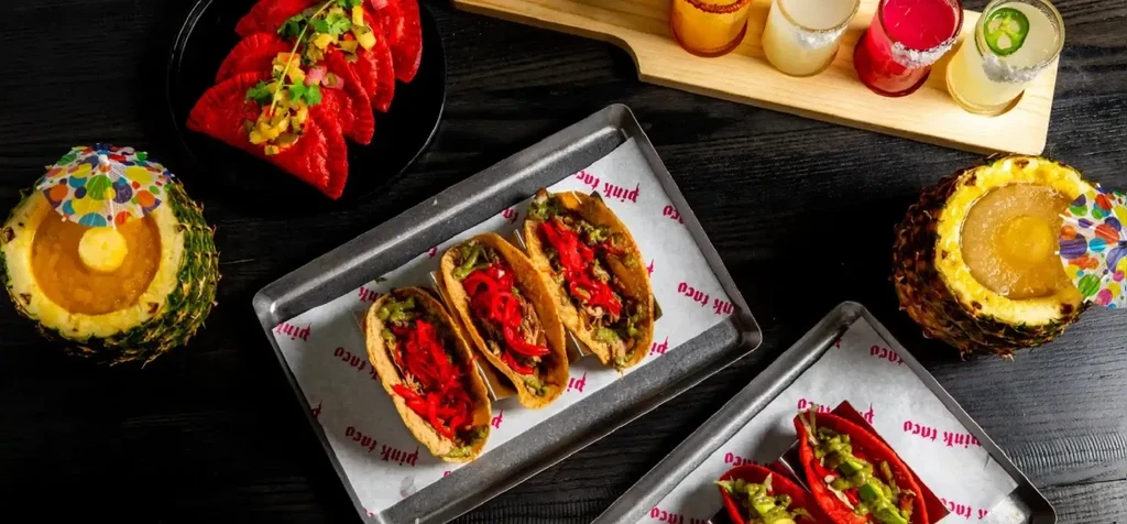 Overhead view of various tacos, colorful drinks, and pineapple cocktails.