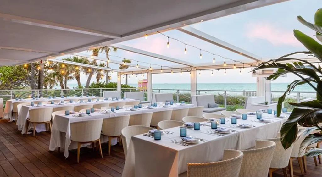 Outdoor dining tables with white linens and blue glasses near a beach.