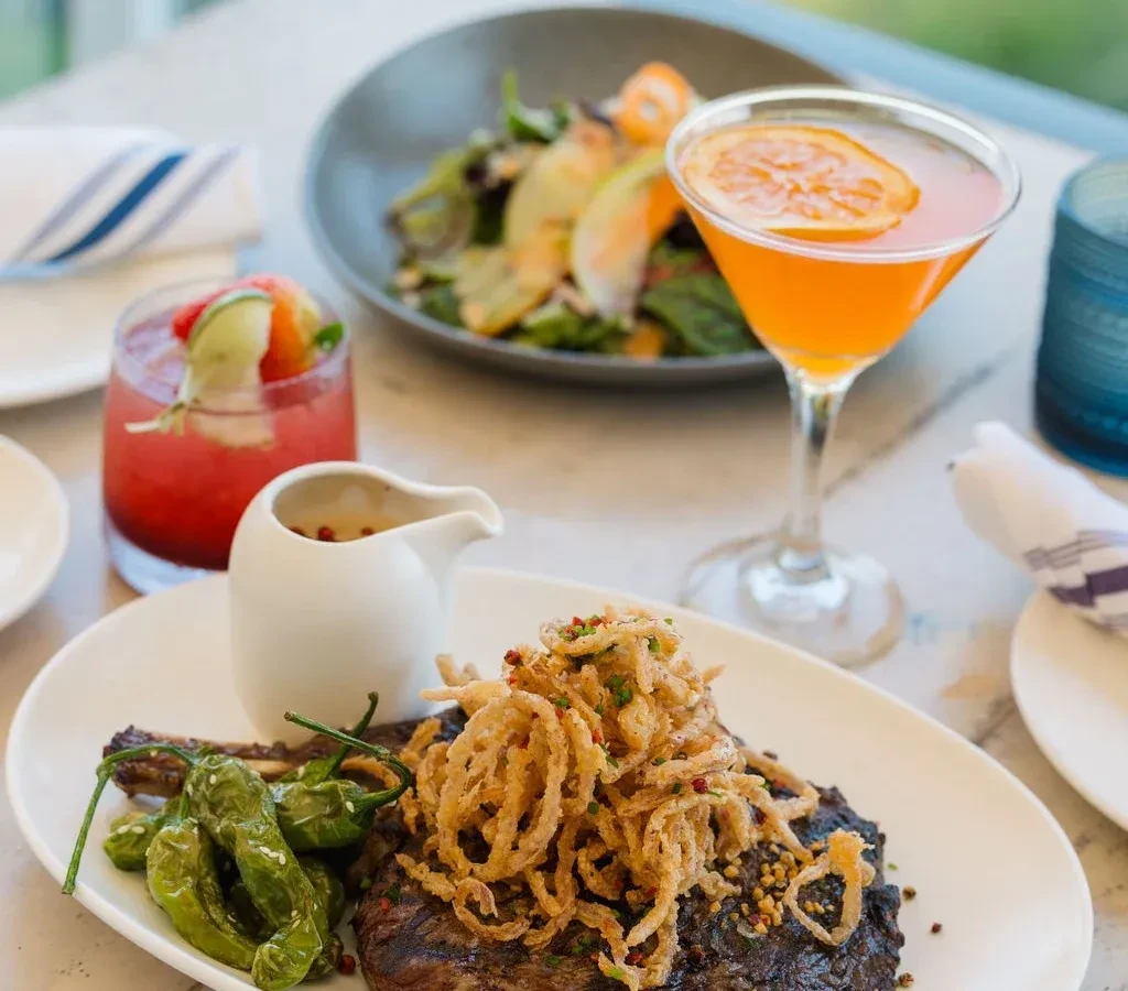 Dining table with grilled steak, onion rings, peppers, and two cocktails.