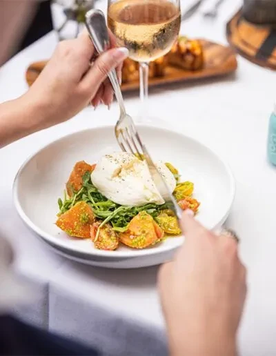 Hands cutting burrata cheese, arugula, and roasted tomatoes.