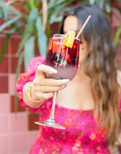Person's hand holds vibrant red-purple cocktail with fruit in stemmed glass.