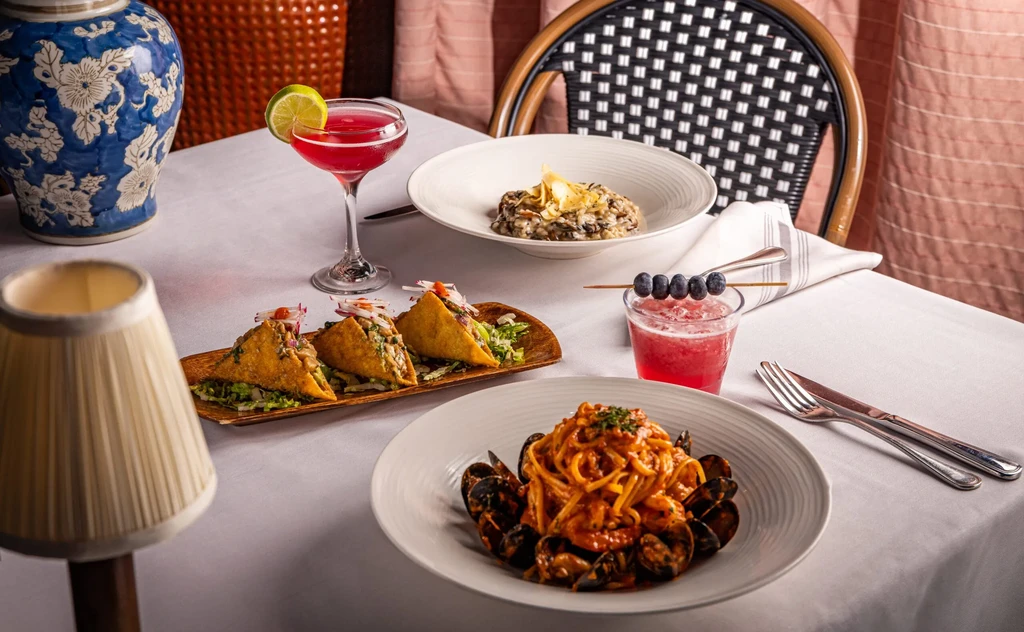 A restaurant table displays tacos, spaghetti, risotto, and vibrant red cocktails.
