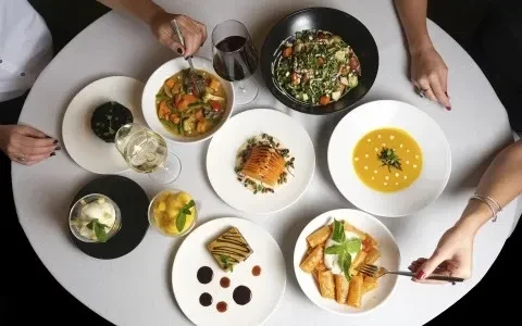 Diners' hands reach for diverse dishes and wines on a round table.