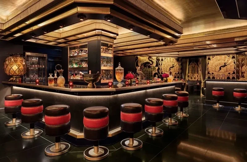 Opulent Art Deco bar with gold bird walls and red-seated stools.