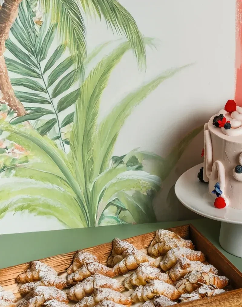 Mini croissants, berry-topped pink cake, and tropical foliage mural.