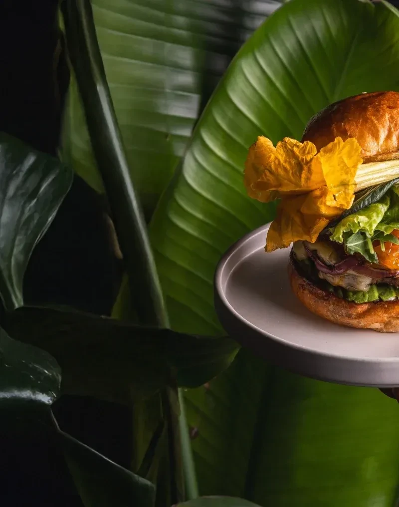 Gourmet burger with edible flower, set against lush tropical leaves.