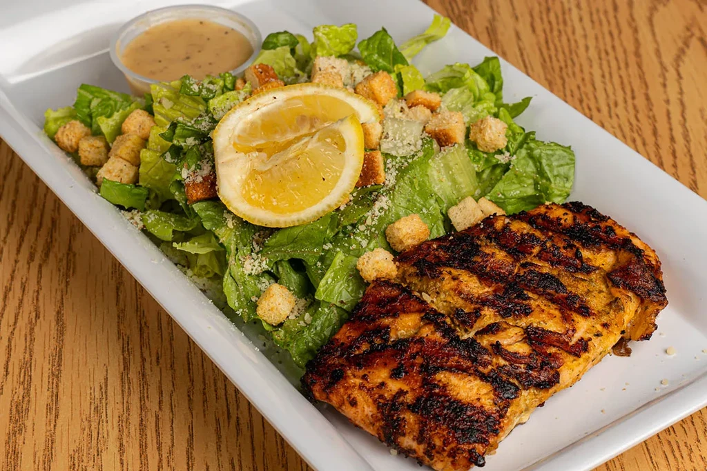 Overhead shot of grilled protein, Caesar salad, and lemon on white plate.