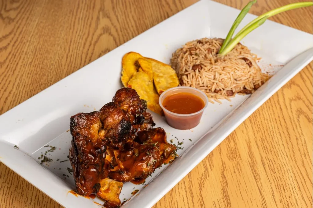 Well-plated glazed ribs, fried plantains, rice, and sauce on wooden table.