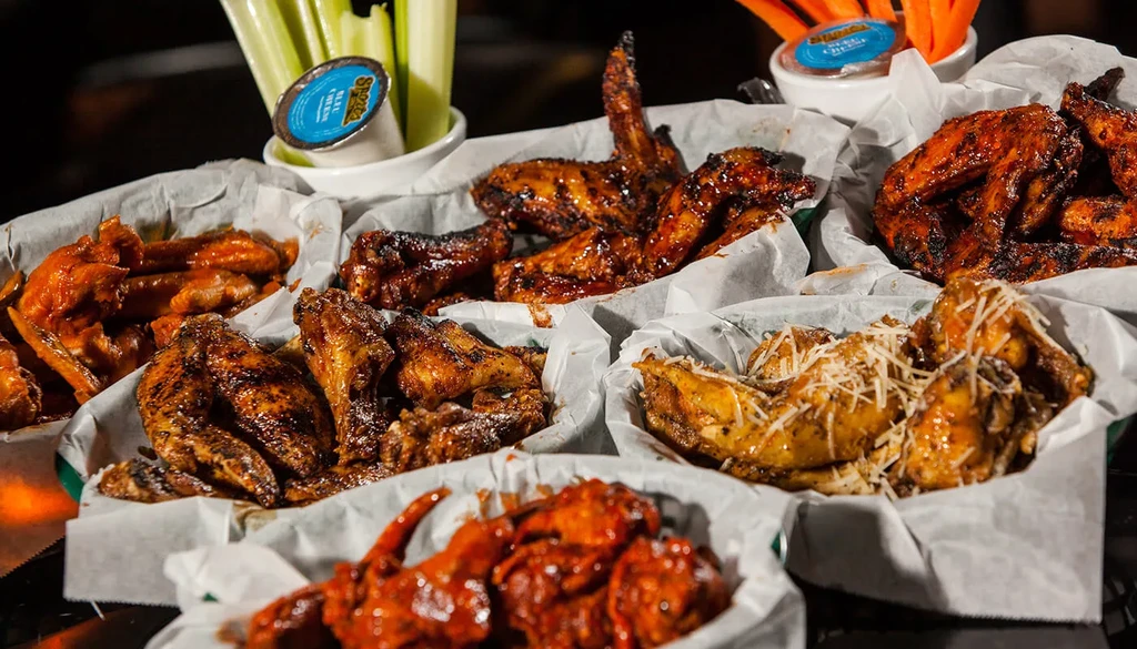 Close-up of various sauced chicken wings with celery, carrots, and dips.