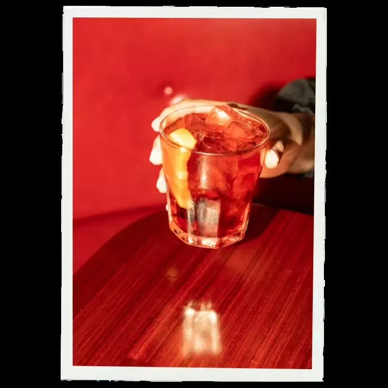 A hand holds a red cocktail with ice and orange peel.