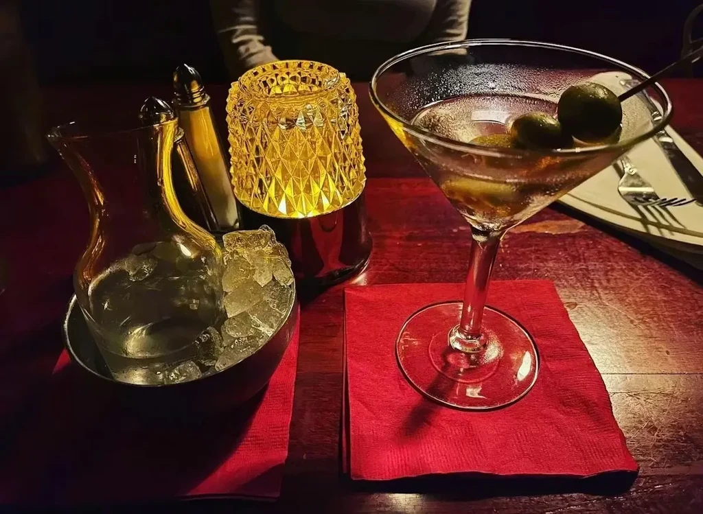 Warmly lit martini, carafe in ice, and lamp on a wooden table.