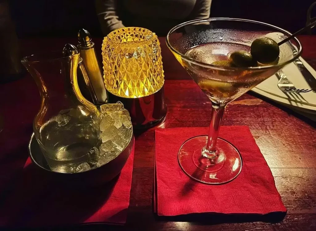 Martini with olives, iced water, and glowing lamp on a table.