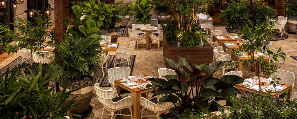 Lush outdoor dining patio with light wood tables and rattan chairs.