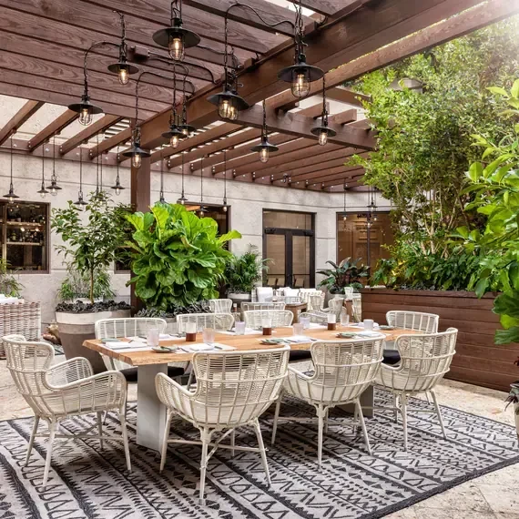 Outdoor dining area with wooden table, white chairs, pergola, and lush plants.