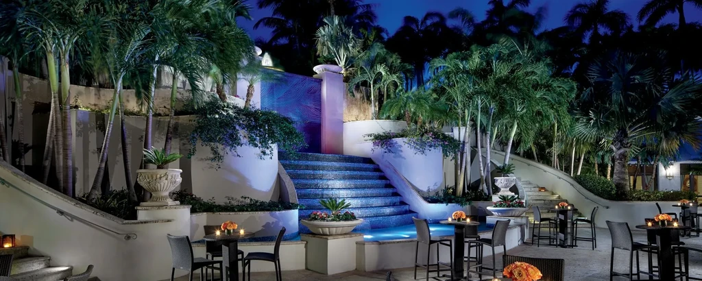 Nighttime outdoor dining with blue-lit water feature and tropical landscaping.