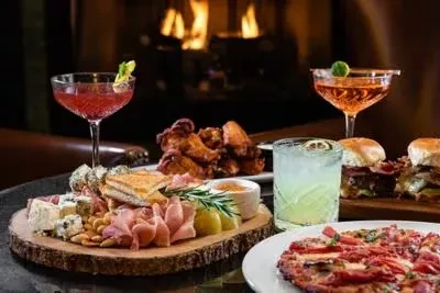 Warm restaurant table with charcuterie, wings, burgers, flatbread, and cocktails.