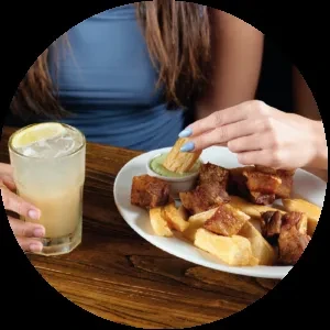 Hands dip fried yuca and chicharrón into sauce, with a drink.