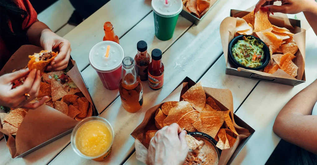 Overhead view of several hands sharing tacos, chips, guacamole, and drinks.