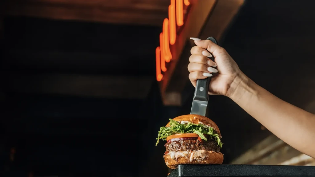 Hand holding knife through a gourmet burger with arugula.