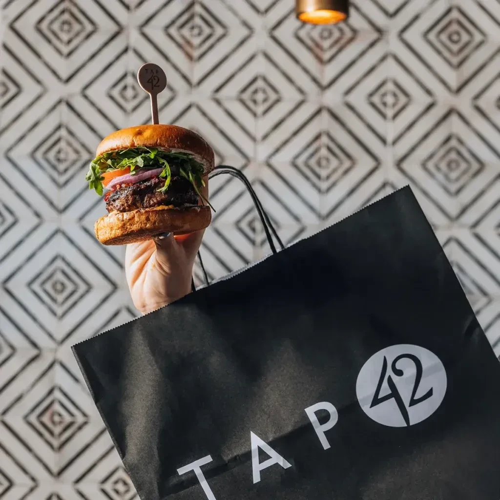 Hand holds a TAP 42 burger above a black branded takeout bag.