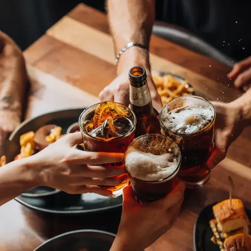 Four hands toasting beer and soft drinks at a wooden table.
