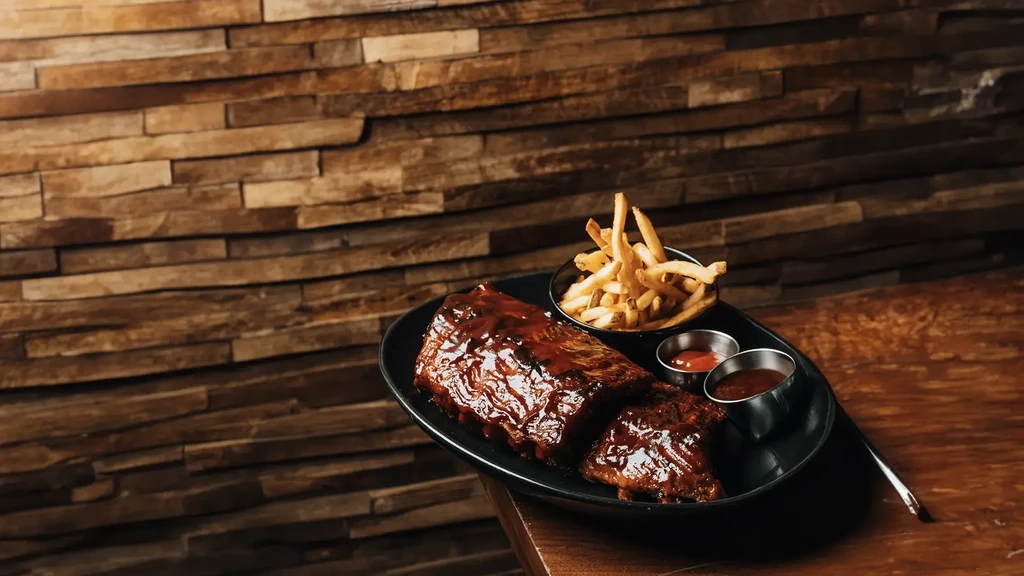 Glistening BBQ ribs, french fries, and two sauces on a black platter.