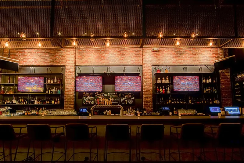 Contemporary bar with stocked shelves, empty stools, and exposed brick walls.