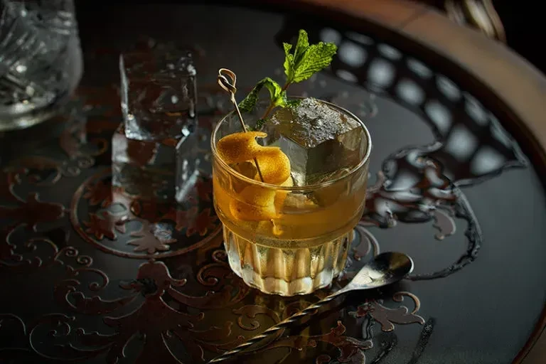 Amber cocktail with ice, orange peel, and mint on a dark ornate tray.