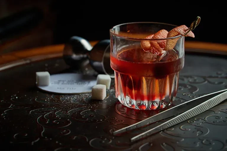 Reddish-amber cocktail with citrus twist, sugar cubes, and bar tools.